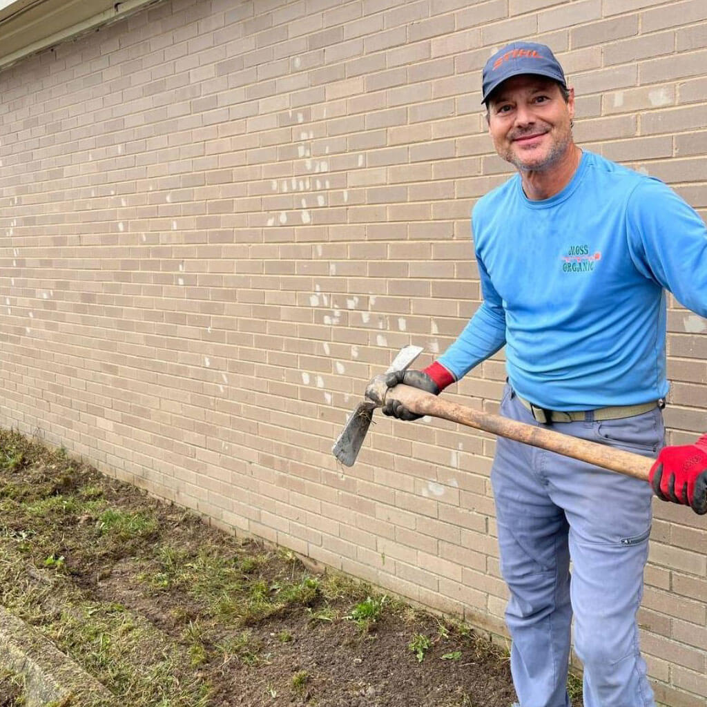 Man in a blue long-sleeve shirt and cap stands with a shovel beside a beige brick wall, smiling while working in a garden bed.