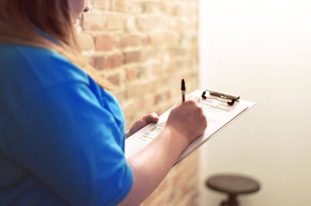 Person in a blue uniform writes on a clipboard labeled 'City Cleaning' beside a brick wall.