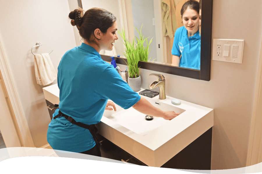Woman in a blue uniform washes her hands at a modern bathroom sink, reflected in the mirror nearby.